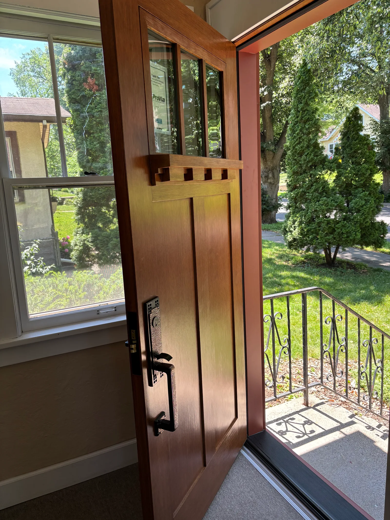 Interior view of a newly installed Craftsman-style front door in stained wood, featuring three vertical glass lites, classic Craftsman detailing, and black mortise lock hardware. The open door looks onto the front porch and a leafy neighborhood beyond.