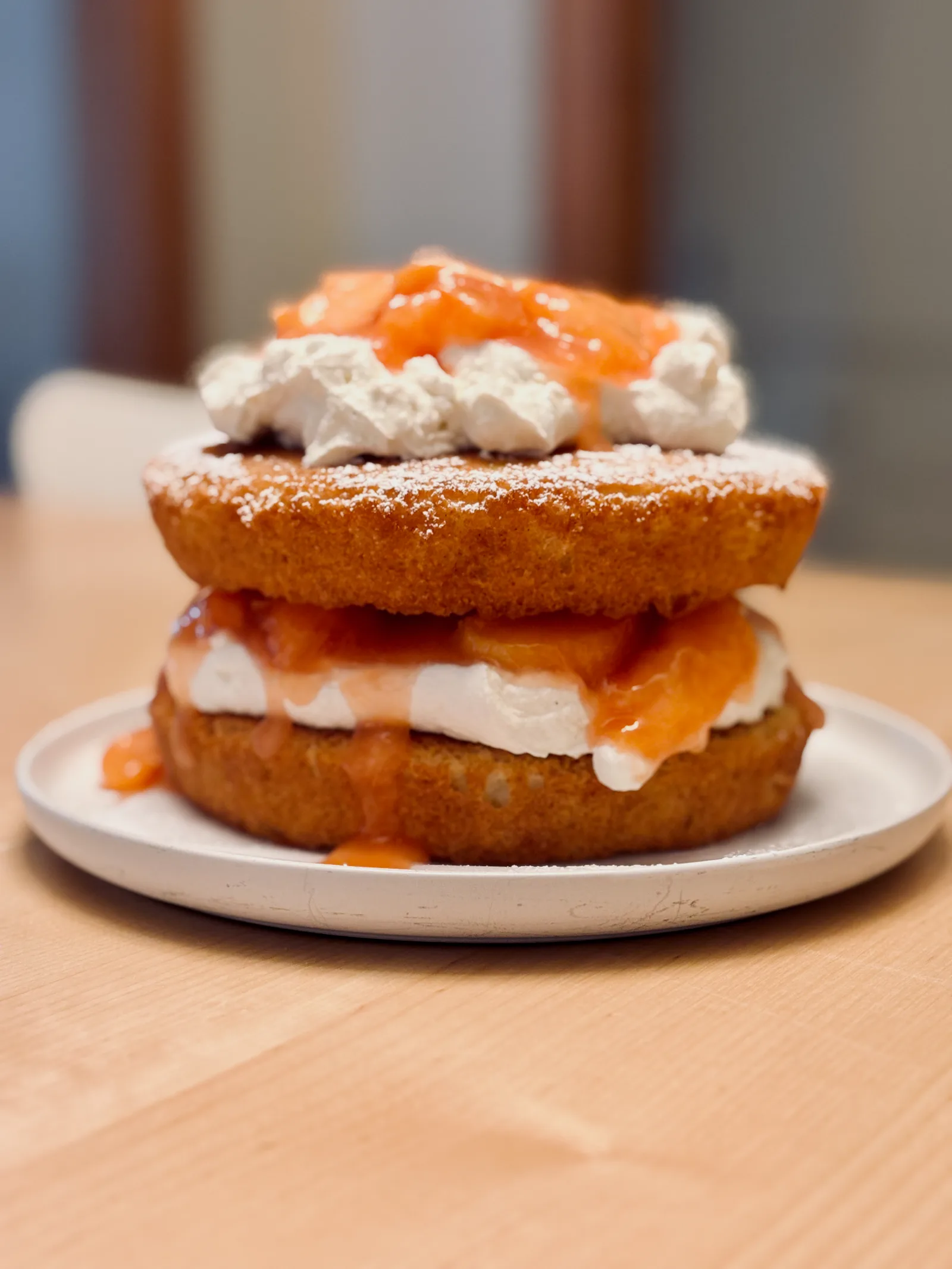 Two-layer sponge cake filled and topped with whipped cream and a glossy orange fruit compote, finished with a dusting of powdered sugar and served on a white plate.